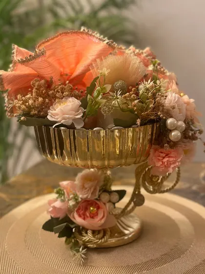 A close up of a golden pedestal bowl filled with beautifully wrapped goodies and decorated with peach and pink flowers. This is a perfect example of our luxurious and detailed work.