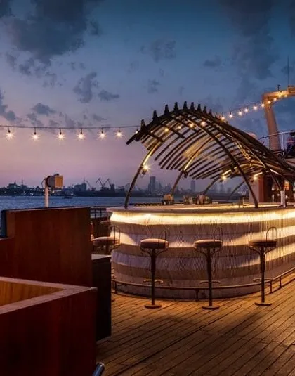 A wider perspective of the upper deck at night. The combination of the sculptural bar, ambient string lights, and the expansive wooden deck creates a perfect setting for events and luxury dining on the water.