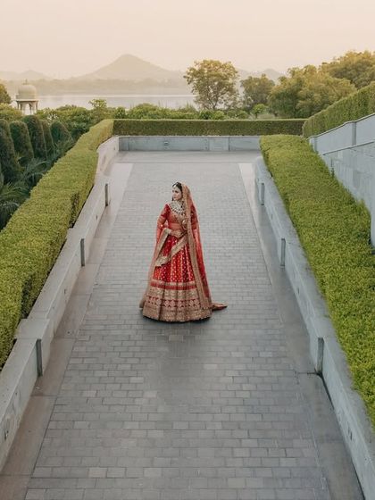 A majestic portrait of Divya at Raffles Udaipur. The wide shot captures her in her stunning red Sabyasachi lehenga against the grand architecture and manicured gardens of the venue.