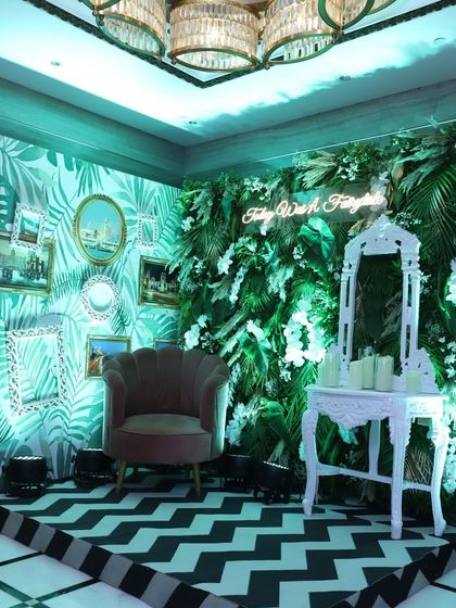 A chic and tropical photo booth. The design features a green leaf-print wall, a vertical garden, vintage frames, and a comfortable armchair, creating a stylish and inviting corner for photos.
