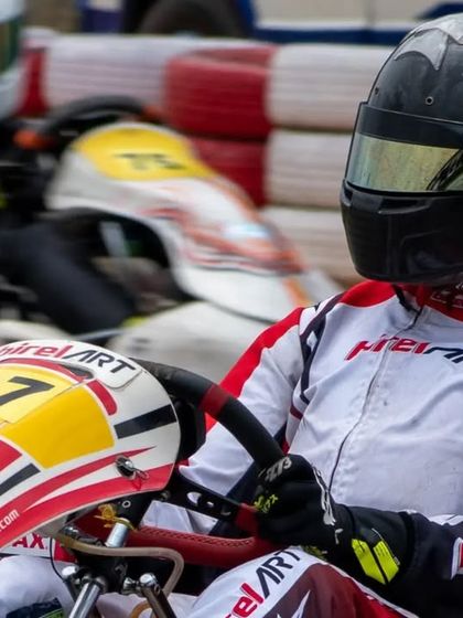 A close-up of driver Muktak Joshi in the cockpit, focused on the race ahead with another kart blurring past in the background.