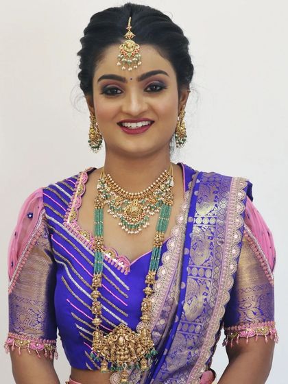A radiant bride ready for her event. Her makeup features a pop of color on the eyes and a bright smile, paired with stunning emerald and gold jewellery that complements her purple and pink silk blouse.