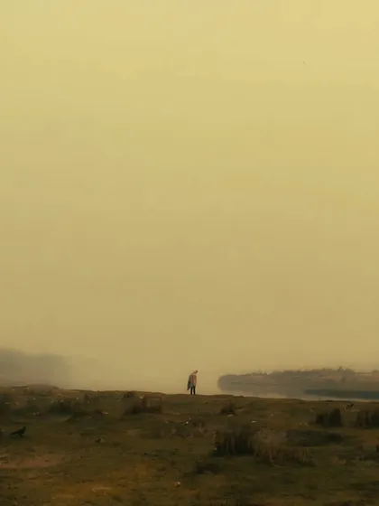 A highly minimalist and moody shot of a lone figure standing in a vast, hazy landscape. The yellow, foggy tones and the smallness of the person create a feeling of isolation and mystery.