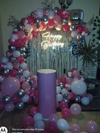 A beautiful birthday backdrop with a pink, silver, and white balloon arch, a neon sign, and a silver fringe curtain.