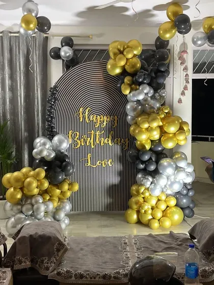 A modern and artistic birthday setup for a loved one. The black, gold, and silver balloon arch is set against a striped backdrop with custom gold lettering.