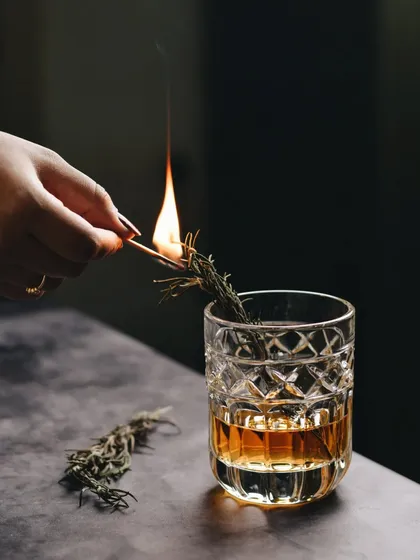 An action shot of lighting a sprig of rosemary for a smoked cocktail. The flame and the dramatic lighting create a sense of theatre and sophistication.