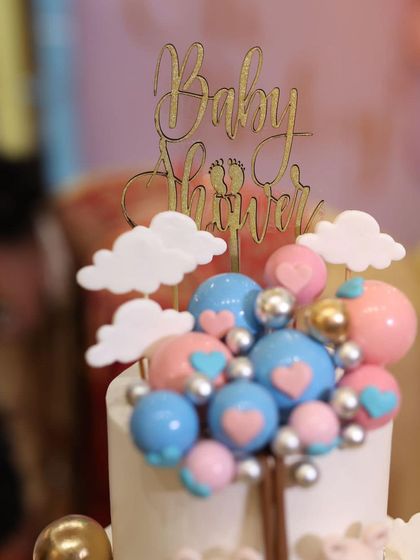 A close-up of the topper for a baby shower cake, featuring a cluster of pink and blue hearts and spheres under a "Baby Shower" sign.