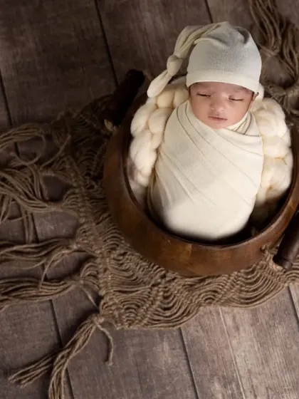 A beautifully composed overhead shot of a swaddled newborn in a wooden bowl. The dark wood floor and rustic macrame mat create a rich, textured look.