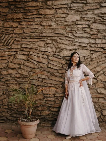 A full-length bridal portrait against a rustic stone wall. The bride's elegant white gown and confident pose create a stunning and timeless image.