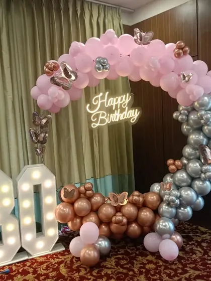A grand 80th birthday celebration with an elegant balloon ring. The pink, rose gold, and silver balloons are accented with butterflies, complemented by a neon sign and large marquee numbers.