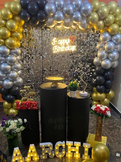 A chic black, gold, and silver themed decoration. The combination of a shimmer wall, balloon arch, and neon sign creates a sophisticated and modern party vibe.