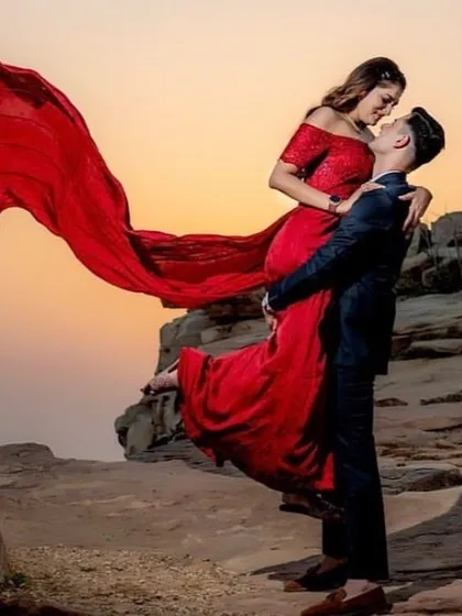 A romantic pre-wedding photo of a couple against a sunset. The bride-to-be is wearing our stunning red off-shoulder gown with a flowing trail.