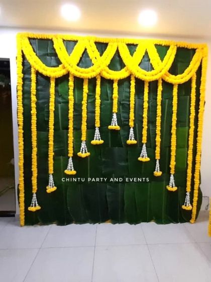 A simple flower decoration for a Half Saree function at home. The backdrop uses a wall of banana leaves and garlands of yellow marigolds.