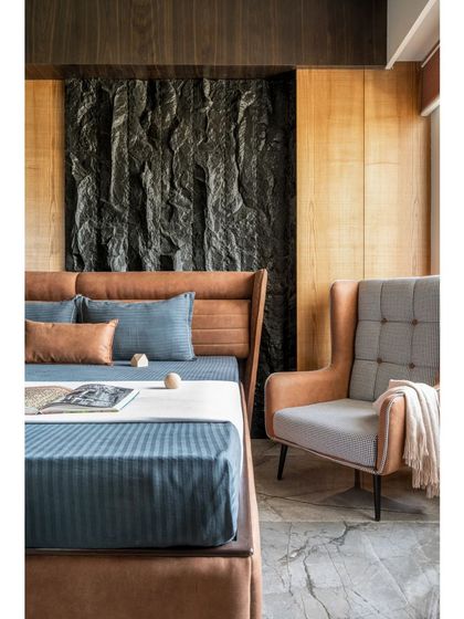 A detail shot of the master bedroom's feature wall and seating nook. The high-back armchair provides a comfortable spot for reading, and its fabric complements the textures of the bed and the natural stone wall.