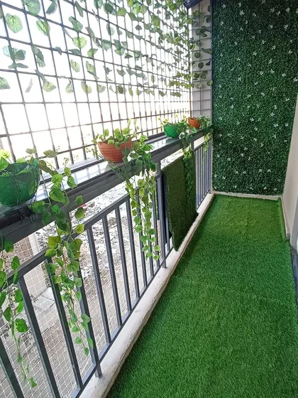 A small apartment balcony made green and private with artificial turf on the floor, an artificial leaf screen on the wall, and hanging plants.