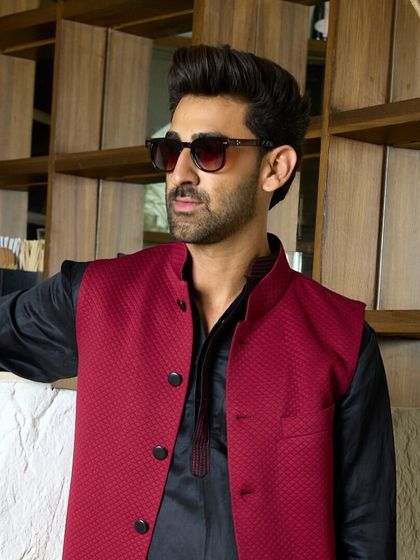 A close-up of the maroon waistcoat, showing the textured fabric. Paired with a black kurta, it's a statement of refined luxury for the modern gentleman.