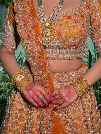 A detail shot focusing on the intricate embroidery of the lehenga and the bride's henna. The overall color palette is warm, bold, and beautifully traditional.