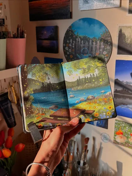 A peaceful gouache painting in my sketchbook, showing a wooden bench by a calm lake surrounded by wildflowers. This is the kind of cottagecore scene I love to create, a perfect spot to sit and dream.