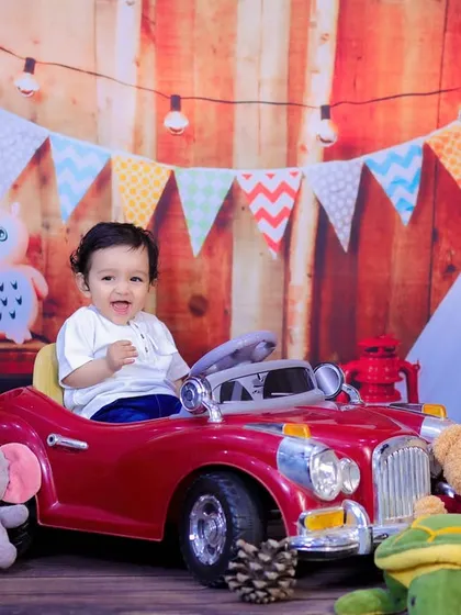 A one-year-old boy in a red toy car, set against our rustic wooden backdrop with colorful bunting. A fun and festive birthday shot.