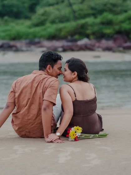 A romantic moment on the beach as the couple sits back-to-back, turning to almost kiss. The flowers beside them add a touch of color to this intimate scene.