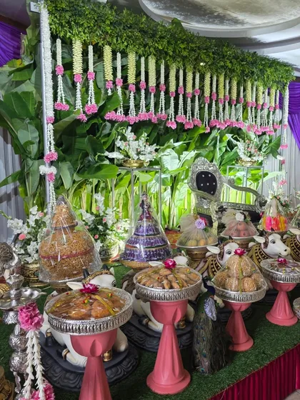 A close-up of an offering table with pink-based cow props and a backdrop of greenery and hanging flowers.