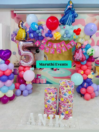A princess party setup with a focus on pink and purple. This decor includes a balloon arch with multiple princess figures, themed plinths, and a silver number "5" balloon.