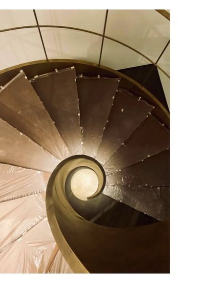 An overhead view of a spiral staircase under construction, its form a perfect curl. Even wrapped in protective plastic, the sculptural quality of the aged brass and wood is evident.