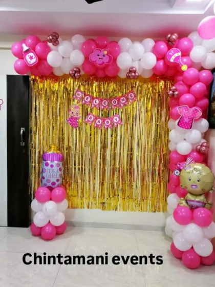 A sweet and simple 'Welcome Baby Girl' decoration for a home. The pink and white balloon arch frames a gold tinsel backdrop, and I've added baby-themed props like a bottle and rattle to complete the look.