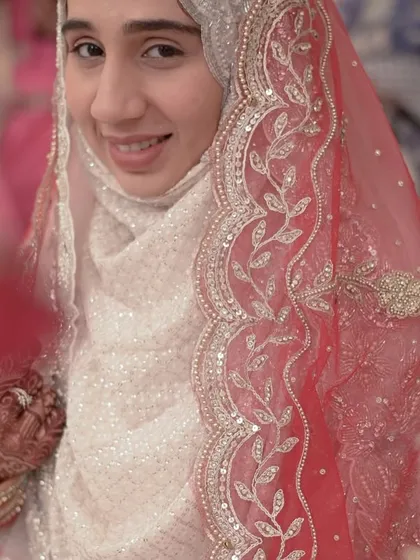A peek through the floral curtain reveals the bride's happy and hopeful smile. This portrait captures the delicate beauty of a hijabi bride on her Nikkah day, framed by traditional decor.