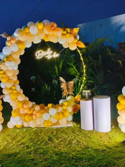 We take the party outdoors with creative setups like this 'Bride to be' celebration. A circular balloon arch in sunset yellows and whites, accented with a neon sign and prop champagne bottle, makes for a perfect garden party backdrop.
