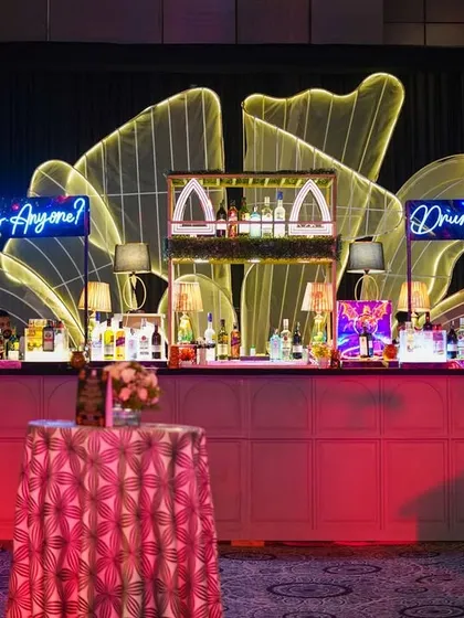 A stunning bar setup for a cocktail night. The bar front is illuminated in pink, with neon signs that say "Cocktails Anyone?" and "Drink in Love," creating a fun and inviting atmosphere for guests.