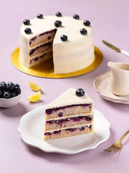 A clean, bright shot of a layered blueberry lemon cake for The Big Chill Cafe. This image is styled for catalog and social media use, emphasizing the cake's structure and fresh ingredients against a simple, colorful background.