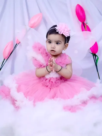 A beautiful baby girl dressed as a fairy with pink wings, sitting among clouds.