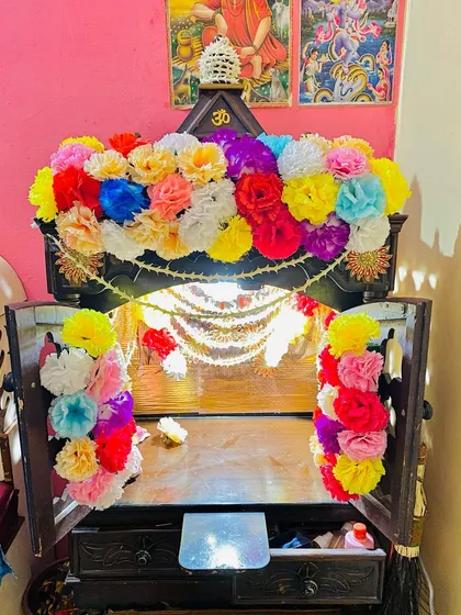 A full view of the wooden home mandir with its doors open. The entrance is decorated with bunches of multi-colored artificial flowers, making it look vibrant and inviting.