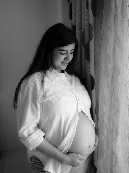 A classic black and white window-lit portrait. The mom-to-be stands by a curtain, the soft light beautifully highlighting her baby bump and serene expression.