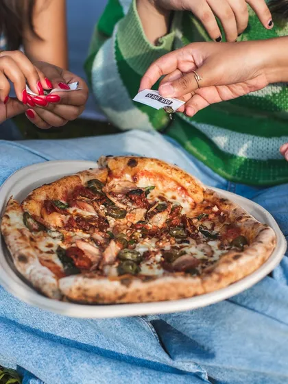 A freshly baked pizza served on a biodegradable plate, ready to be shared. Notice the perfectly charred and bubbly sourdough crust, a hallmark of our baking process.