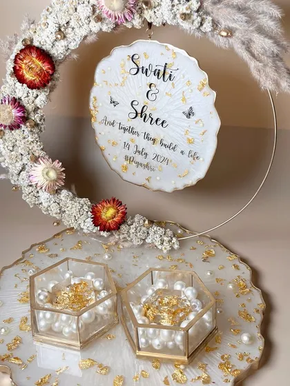 An elegant white and gold ring platter with a floral hoop featuring dried flowers and pampas grass. The hexagonal ring boxes are filled with pearls and gold flakes.
