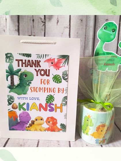 A complete dinosaur-themed return gift, showing the personalized mug wrapped in netting and the matching custom paper bag.