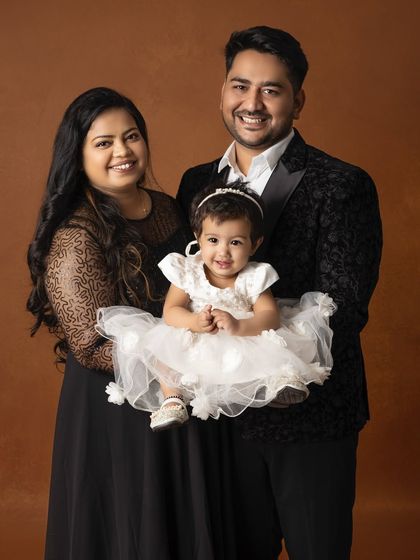 A lovely portrait of a young family. The parents hold their daughter, who is dressed in a beautiful white dress, all sharing warm smiles.