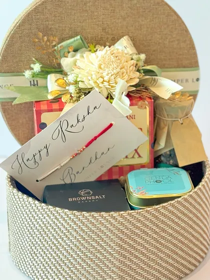 An inside look at the round woven Rakhi box, showcasing the carefully packed trail mix, artisanal soap, and other little surprises. The floral arrangement on top makes it feel extra special.