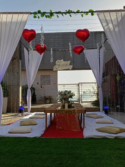 A romantic rooftop sit-down dinner setup for a birthday. The canopy is draped with white curtains and decorated with heart-shaped balloons and fairy lights.