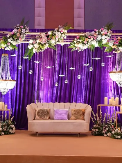A royal reception stage with a backdrop of rich purple velvet, accented with mini chandeliers and framed by an arch of beautiful white and pink flowers.