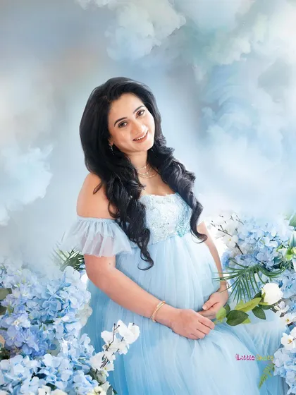 A close-up from a fantasy-themed shoot, where the mother-to-be is surrounded by blue flowers. She holds a single white rose, smiling gently in her ethereal blue gown.