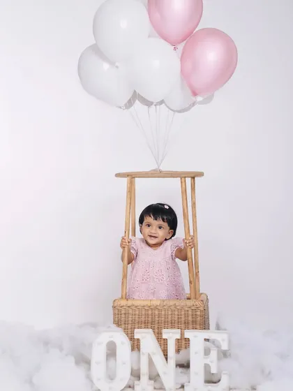A dreamy and classic first birthday studio portrait. The hot-air balloon basket, cloud-like floor, and soft pink balloons create a whimsical atmosphere perfect for celebrating your baby's special day.