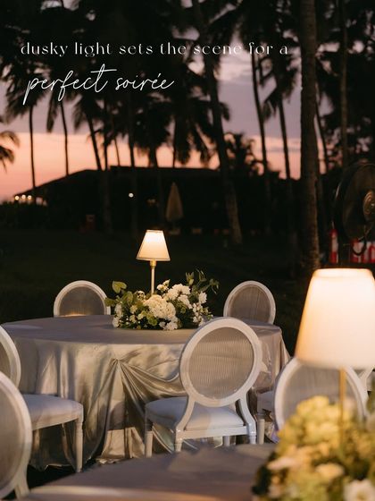 A table at the Goa soirée, with a beautiful floral centerpiece and elegant lamp lighting, with the text overlay "dusky light sets the scene for a perfect soirée".