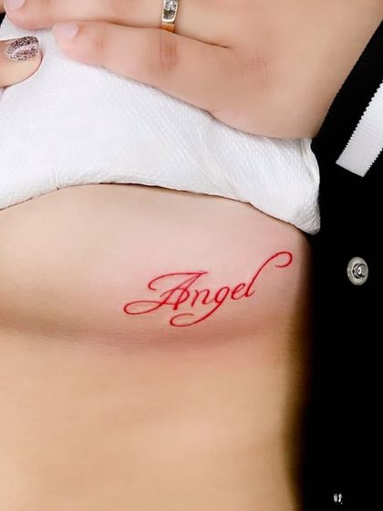 An "Angel" script tattoo in red ink on the rib cage.