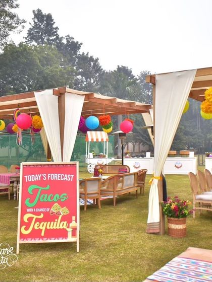 A fun sign reading 'Today's Forecast: Tacos with a chance of Tequila' adds a playful touch to this outdoor fiesta setup, which includes pergolas and picnic-style seating.