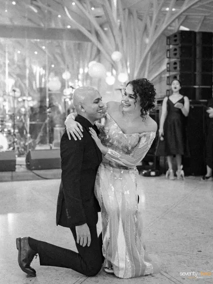 A joyful, candid moment of the groom on one knee before his bride on the dance floor, both of them laughing.