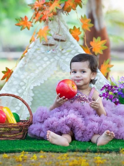 A sweet toddler in a fluffy purple dress sits in front of a teepee, holding an apple with a curious smile. The colorful props and natural setting make for a magical portrait.
