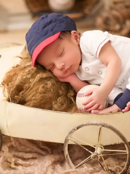 A future baseball star. This baby is dressed in a mini baseball uniform, sleeping in a vintage-style toy car and holding a baseball.